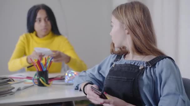 Cute biały nastolatek siedzi ze smartfonem jako jej Afroamerykanin przyjaciel daje jej kartkę papieru. Przyjaciele spędzają razem czas w domu. Styl życia, edukacja, nauka. — Wideo stockowe