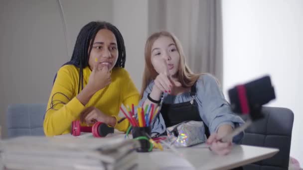 Portræt af to venner gør videoblog sammen. Kaukasiske og afroamerikanske teenagepiger kigger på smartphone skærm og taler. Bedste venner hviler sammen indendørs . – Stock-video