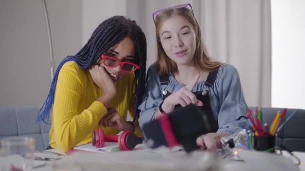 Positiva tonårsvänner som pratar med selfiekamera och skrattar. Vit tjej och hennes afroamerikanska vän i röda glasögon har kul inomhus. Lycka, livsstil. — Stockvideo