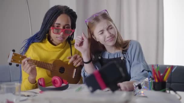 Chica afroamericana jugando ukelele como su amiga caucásica bailando. Adolescentes positivos que se divierten como grabación de vídeo selfie. Felicidad, estilo de vida, descanso . — Vídeos de Stock