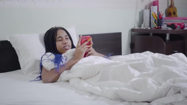 Câmera aproximando menina afro-americana com dreadlocks deitado na cama branca e usando smartphone. Jovem surfando na Internet à noite. Vício em mídias sociais, estilo de vida . — Vídeo de Stock