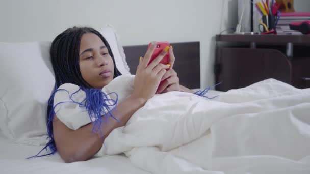 Caméra approchant fille afro-américaine avec dreadlocks ranger smartphone et regarder vers le haut réfléchie. Adolescent utilisant les médias sociaux au coucher. Dépendance à Internet, mode de vie . — Video