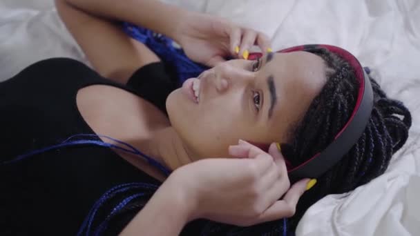 Zbliżenie twarzy Afroamerykanki z dredami leżącej na białym łóżku i słuchającej muzyki w słuchawkach. Nastolatek bierze smartfona i zmienia piosenkę. Wypoczynek, dojrzewanie, styl życia. — Wideo stockowe