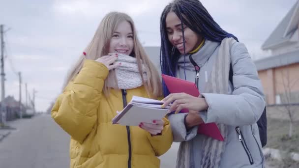 Egy afro-amerikai lány portréja, amint kaukázusi barátjával beszél, és átnézi a jegyzeteit. Pozitív diákok állnak az utcán és beszélgetnek. Boldogság, életstílus. oktatás. — Stock videók