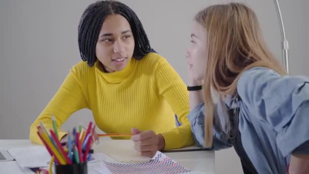 アフリカ系アメリカ人の友人と話している白人の少女の側面図と感情的にジェスチャー。友達は屋内で宿題をしながら話す。生活、教育、幸福. — ストック動画