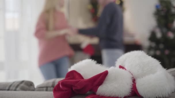 Gros plan de chapeaux de Noël rouges couchés sur le canapé comme des gens caucasiens heureux dansant en arrière-plan. Un jeune couple positif qui s'amuse la veille du Nouvel An à la maison. Vacances, loisirs, mode de vie . — Video