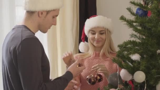 Hermosa mujer caucásica en sombrero de Navidad dando decoraciones al hombre adulto colgándolos en el árbol de Año Nuevo. Feliz pareja decorando la casa para las vacaciones. Felicidad, ocio, celebración . — Vídeos de Stock