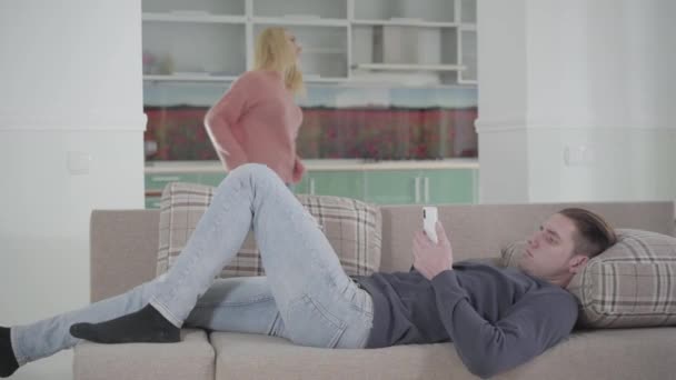 Portrait of young Caucasian man lying on sofa and using smartphone while his unhappy wife vacuuming at the background. Man ignoring household issues. Relationship problems. — Stock Video