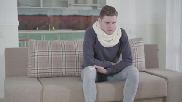 Portrait of Caucasian young man in white scarf sitting on sofa and coughing. Sick guy having sore throat. Illness, healthcare and medicine. — Stock Video