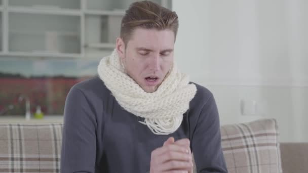 Close-up of Caucasian man in white scarf sitting on sofa and sneezing. Portrait of sick guy at home. Illness, healthcare and medicine, family care. — Stock Video