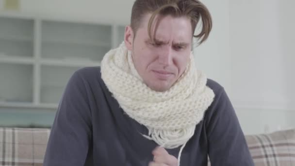 Close-up portrait of Caucasian man in white scarf sitting on sofa, sneezing and coughing. Portrait of sick guy shivering at home. Illness, healthcare and medicine. — Stock Video