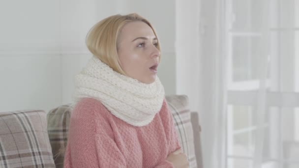 Retrato de una joven atractiva mujer caucásica en pañuelo blanco temblando. Chica rubia sentada en casa en el sofá y estornudando. Salud, enfermedades médicas, enfermedades infecciosas . — Vídeos de Stock