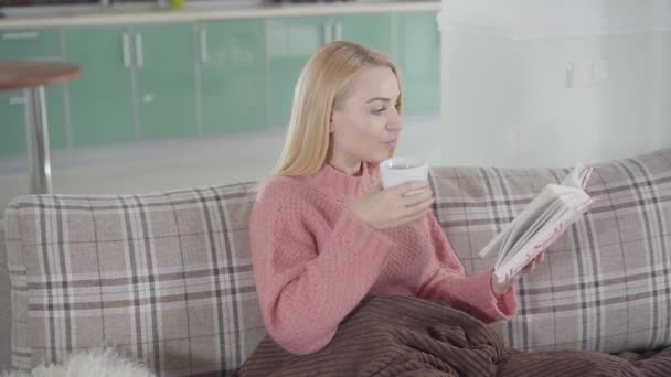 Porträtt av charmig vit kvinna sitter med bok och kopp varmt te eller kaffe hemma. Söt flicka läser intressant historia, vänder blad och dricka ur koppen. hobby, fritid, livsstil. — Stockvideo