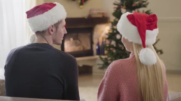 Back view of young happy Caucasian couple sitting in front of Christmas tree. Woman in red Christmas hat putting head on husbands shoulder. Holidays season, love, relationship. — Stock Video