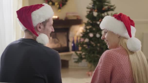 Achteraanzicht van positief Kaukasisch koppel dat lacht als zittend voor de kerstboom. Vrouw en man met rode kerstmutsen hebben plezier op oudejaarsavond. Vakantie seizoen, liefde, relatie. — Stockvideo