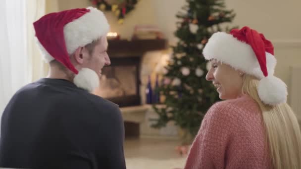 Vista trasera de pareja caucásica positiva riendo como sentado frente al árbol de Navidad. Mujer y hombre con sombreros rojos de Navidad divirtiéndose en Nochevieja. Vacaciones temporada, amor, relación . — Vídeos de Stock