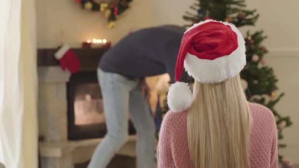 Achteraanzicht van een blonde blanke vrouw zittend voor de kerstboom als jonge man die een geschenkdoos oppakt. Vrouw of vriendin wachtend op echtgenoot om haar cadeau te geven op oudejaarsavond. — Stockvideo