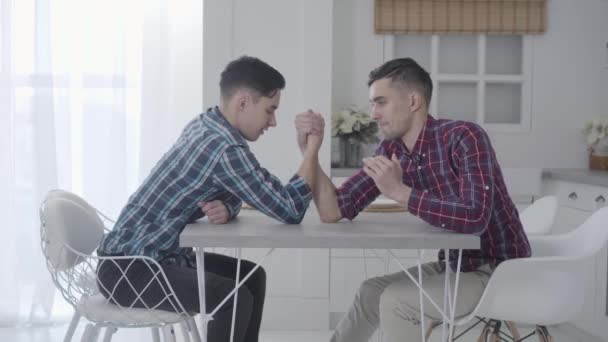 Hermanos gemelos caucásicos compitiendo en armwrestling en casa. Dos morenos adultos positivos sentados a la mesa en el interior. Diversión, ocio . — Vídeos de Stock