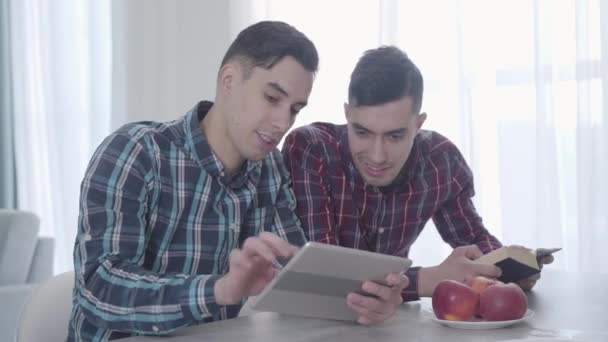 Portrait d'un homme caucasien adulte jouant à un jeu vidéo sur tablette et le montrant à son frère jumeau. Brunette gars lecture livre et regarder l'écran de l'appareil. Frères et sœurs qui reposent à l'intérieur . — Video