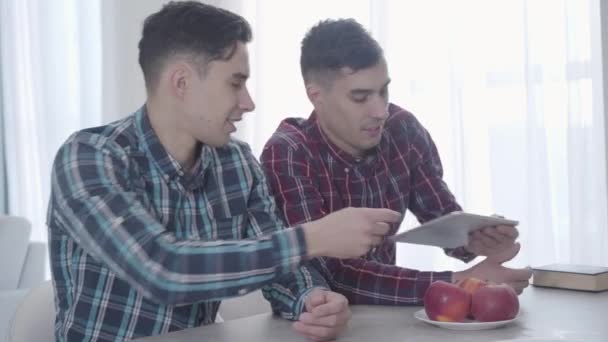 Volwassen blanke man geeft tablet aan zijn tweelingbroer, broer en zus geeft hem boek. Twee identieke tweelingen rusten in het weekend thuis. Vrije tijd, geluk, levensstijl. — Stockvideo