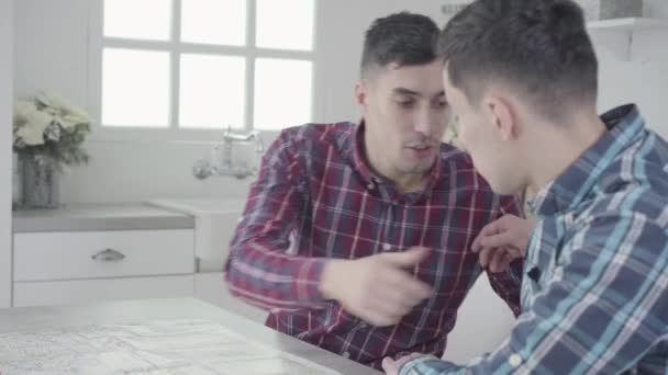 Retrato de hermanos gemelos idénticos discutiendo sobre el atuendo. Hermanos tocándose camisas casuales y hablando. Hermanos preparándose para un viaje, mapa acostado en la mesa. Estilo de vida, turismo . — Vídeos de Stock