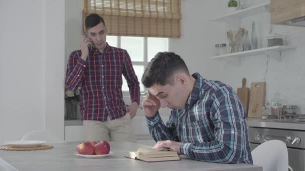 Lelaki Kaukasia yang kesal duduk di meja dan menggosok kuil sebagai saudara kembarnya berbicara di telepon pintar di latar belakang. Guy mengganggu saudaranya dari membaca. Kesenangan, gaya hidup . — Stok Video
