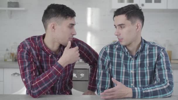 Close-up of two adult Caucasian twin brothers talking to each other, turning to camera and smiling. Positive young men posing at home. Friendship, family, identical siblings. — Stock Video