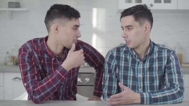 Primer plano de dos hermanos gemelos caucásicos adultos hablando entre sí, girando hacia la cámara y sonriendo. Jóvenes positivos posando en casa. Amistad, familia, hermanos idénticos . — Vídeos de Stock