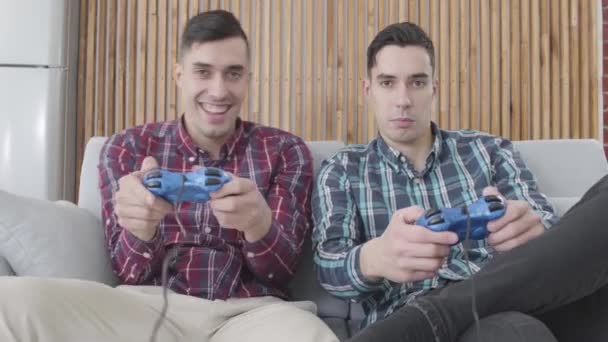 Two positive Caucasian men competing in video game. Guy in red shirt showing finger on his twin brother when he looses and laughing. Men having fun on weekends at home. Leisure, happiness, resting. — Stock Video