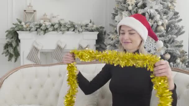 Retrato de menina branca bonita feliz em chapéu de Natal vermelho colocando em guirlanda amarela, olhando para trás na árvore de Ano Novo decorado, e sorrindo. Mulher alegre se preparando para as férias. Estilo de vida . — Vídeo de Stock