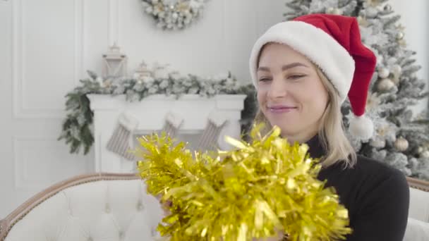 Primer plano de la hermosa mujer caucásica en sombrero rojo que se pone la guirnalda amarilla, mirando hacia atrás en el árbol de Año Nuevo decorado, y sonriendo. Chica feliz preparándose para las vacaciones. Estilo de vida . — Vídeos de Stock