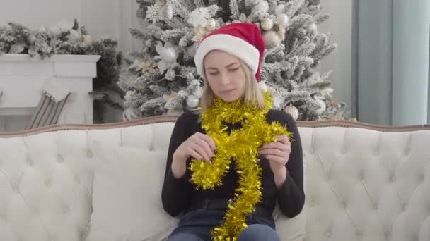 Retrato de una atractiva mujer caucásica sentada a la luz del sol en el sofá, chica triste esperando a los invitados o amigos en el fondo del árbol de Navidad en la víspera de Año Nuevo. Vacaciones temporada, celebración . — Vídeo de stock