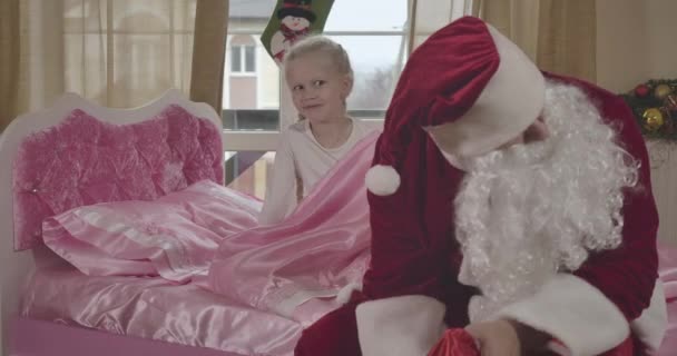 Surpris fille caucasienne assise sur un lit rose et souriant comme le Père Noël en costume rouge regardant dans le sac-cadeau. Enfant ayant miracle la veille de Noël à la maison. Siège social Cinema 4k ProRes . — Video