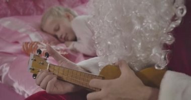 Arka planda uyanan küçük sarışın kız olarak Noel Baba 'nın ukulele çaldığı yakın plan. Baba pembe odada sevgili kızını uyandırıyor. Tatiller, Noel, yeni yıl. Sinema 4k Prores Merkezi.