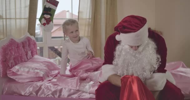 Beyaz adam portresi Noel Baba kostümü giymiş pembe yatağın üzerinde oturan at kuyruklu küçük kız omzuna dokunuyor. Noel sabahı evde uyanan bir çocuk. Sinema 4k Prores Merkezi. — Stok video
