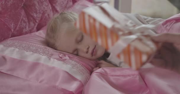 Portret pięknej białej dziewczynki śpiącej w różowym łóżku jako męska dłoń kładąca prezent na poduszce. Sezon wakacyjny, cud, szczęście, dzieciństwo. Kino 4k Prores Hq. — Wideo stockowe