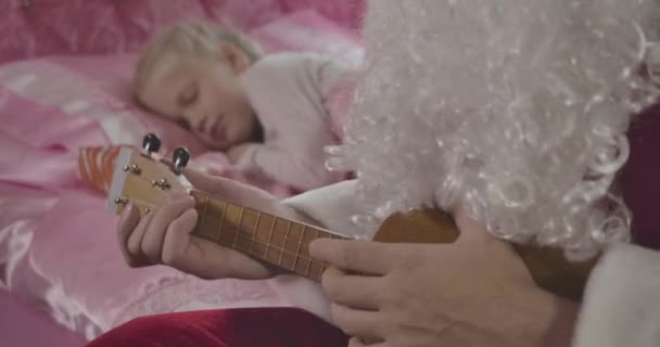 Close-up of unrecognizable Caucasian man in Santa Claus costume with false beard playing ukulele as little blond girl sleeping at the background. Cinema 4k ProRes HQ. — Stock Video