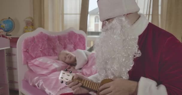 Zamazana biała dziewczyna budzi się rano i widzi Świętego Mikołaja siedzącego na różowym łóżku i grającego na ukulele. Ojciec zaskakujący córka w Wigilię. Kino 4k Prores Hq. — Wideo stockowe