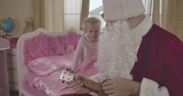 Aranyos fehér gyerek ül a rózsaszín ágyon, és nézi a Mikulás játszik ukulele. Felnőtt férfi Mikulás jelmezben hangszert ad neki. Ünnepek, csoda, boldogság. Mozi 4k Prores Hq. — Stock videók