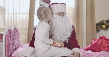 Şok olmuş Noel Baba 'nın portresi güzel beyaz kızın dileklerini dinliyor. Tatlı çocuk Noel Baba 'nın kulağına fısıldıyor ve gülümsüyor. Pembe yatakta oturan bir adam ve çocuk. Sinema 4k Prores Merkezi.