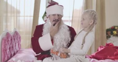 Kafkasyalı kızın Noel Baba kostümlü babasına bakmasına şaşırdım. Takma sakal çıkarılmış, adam yüzüne dokunuyor. Baba ve kızı Noel arifesinde evde eğleniyorlar. Sinema 4k Prores Merkezi.