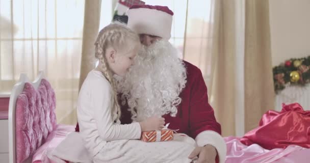 Roztomilá běloška s copánky na kolenou Santa Clause a šeptá mu do ucha. Dítě, které drží přítomnost a sdílí tajemství. Vánoce, svátky, Silvestr. Cinema 4k Prores Hq. — Stock video