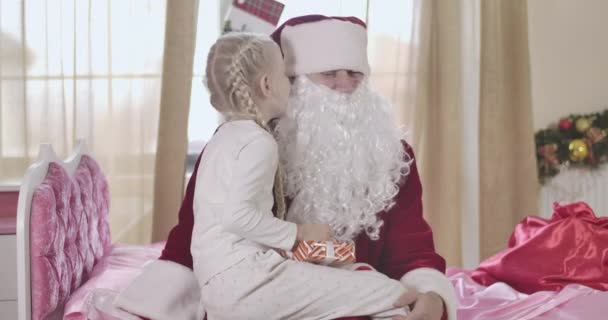 Portrait of shocked Santa Claus listening to wishes of pretty Caucasian girl. Cute child whispering on Santas ear and smiling. Man and kid sitting on pink bed indoors. Cinema 4k ProRes HQ. — Stock Video