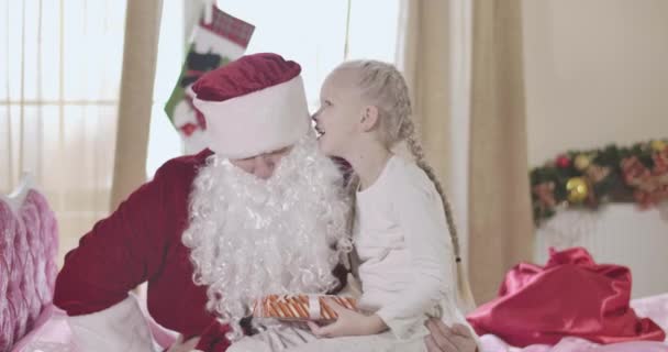 Noel Baba 'nın dizlerine çöküp kulağına fısıldayan neşeli sarışın beyaz kız. At kuyruklu güzel çocuk hediyesini tutuyor ve Noel Baba 'yla konuşuyor. Noel, tatiller. Sinema 4k Prores Merkezi. — Stok video