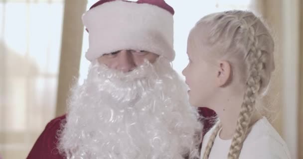 Close-up face of cute blond Caucasian girl with pigtails whispering on Santas ear. Pretty kid talking to Santa indoors. Christmas, New Year, holidays. Cinema 4k ProRes HQ. — Stock Video