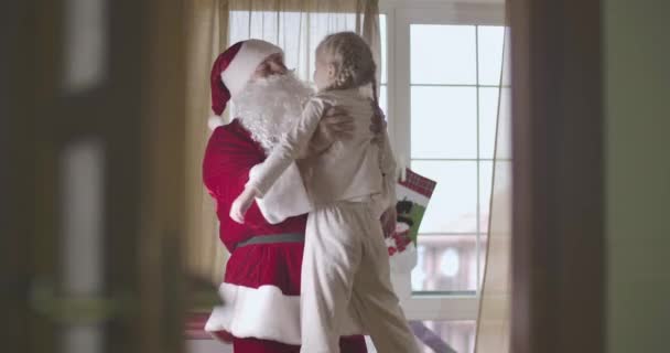 Hombre caucásico en traje de Papá Noel girando niño caucásico bastante rubio. Chica sonriente con coletas en pijama divirtiéndose con Santa en la víspera de Navidad. Sede del cine 4k ProRes . — Vídeos de Stock