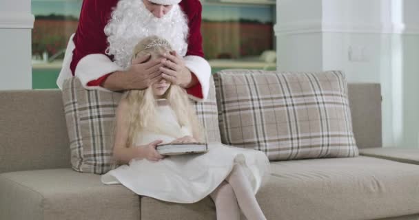 Portrait de fille blanche blonde en robe blanche élégante et couronne assise sur le coach avec les yeux fermés par les mains du Père Noël. Un enfant qui tourne le dos. Siège social Cinema 4k ProRes . — Video