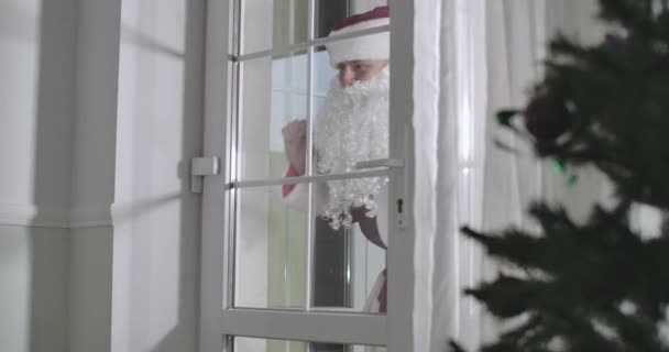 Santa Claus de pie detrás de la puerta de entrada de cristal y saludando como un niño lindo corriendo hacia arriba y saludando hacia atrás. Hombre sonriente con bolsa de regalo que viene a la hermosa chica caucásica. Sede del cine 4k ProRes . — Vídeo de stock