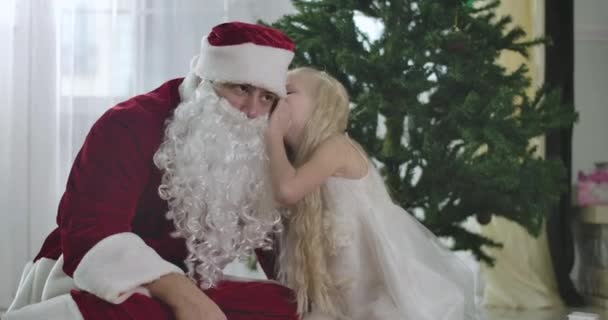 Pretty Caucasian girl sitting with Santa in front of Christmas tree at home and whispering on his ear. Child asking Santa Claus about presents, Cinema 4k ProRes HQ. — Stock Video