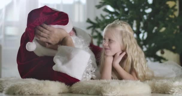 Cute Caucasian girl with blond curly hair lying with Santa at the background of Christmas tree and smiling. Pretty child talking to Santa at home. Cinema 4k ProRes HQ. — Stock Video